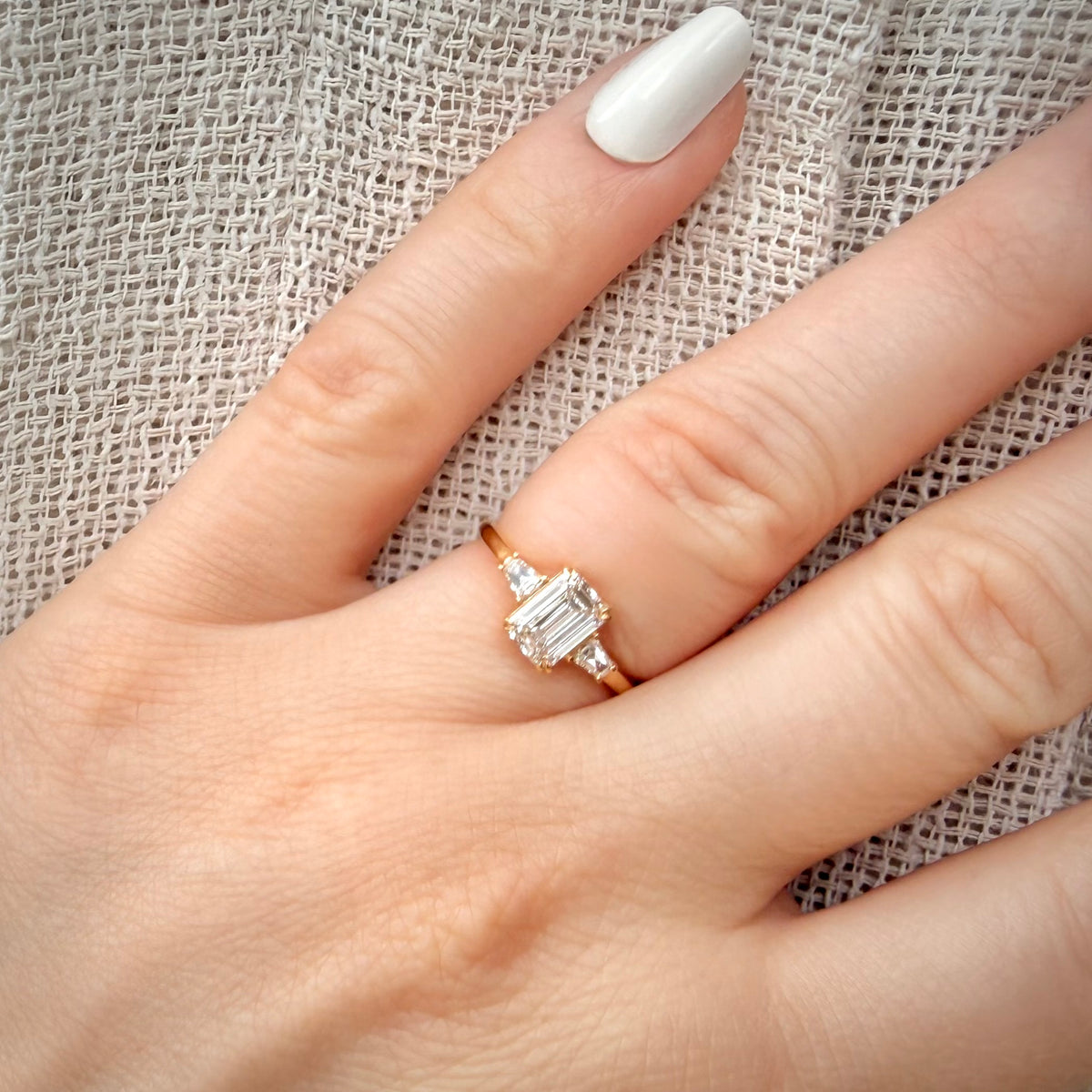 1,20 ct Emerald Cut trilogy-timanttisormus sormessa pellava taustalla