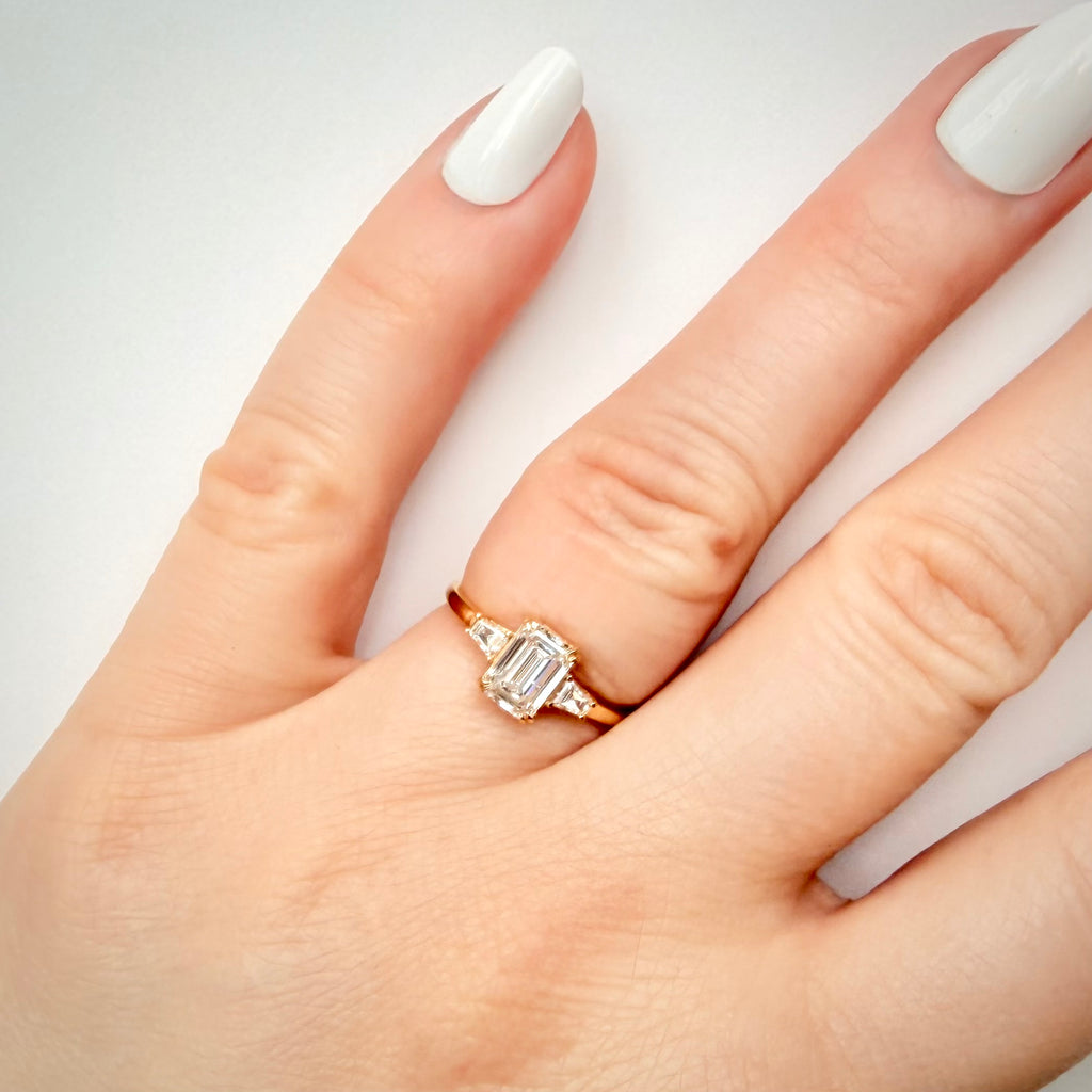 1,20 ct Emerald Cut trilogy-timanttisormus sormessa valkoisella taustalla